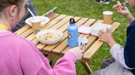 Lapset nauttivat lounasta yhdessä ulkona piknik-pöydän ääressä. Sinisessä juomapullossa on pingviinitarra.