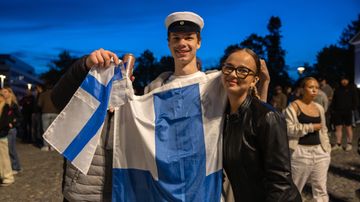 Ylioppilaslakki päässä seisova nuori mies hymyilee kameralle kädessään iso Suomen lippu. Vieressä nuori nainen ja toinen mies, jonka kasvot eivät näy.