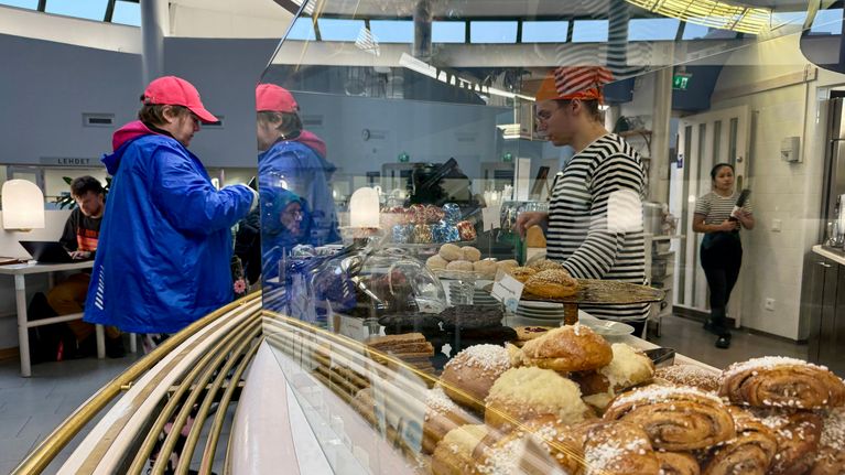 Lähikuvassa Cafe Metson pullatarjontaa. Taustalla asiakas maksamassa ostoksia kahvilan kassalla.