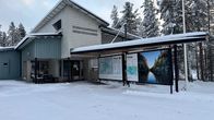 A blue and white wooden building with maps and information boards in front, surrounded by snow and trees.