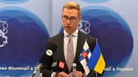 En man i kostym står vid ett podium med mikrofoner och finska samt ukrainska flaggor, framför en blå bakgrund med texten "Embassy of Finland in Ukraine".