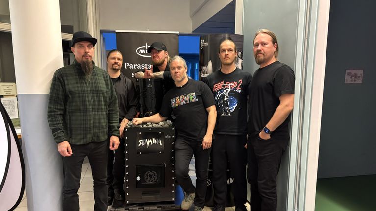 Kuusi miestä seisoo sisätiloissa saunakiukaan ympärillä.