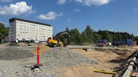 Rakennustyömaa kesällä, sepeliä etualalla, kaivinkone vasemmalla, taustalla sininen sorarekka. 