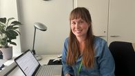 Nainen istuu toimistossa, katsoo kameraan ja hymyilee. Pöydällä läppäri, lamppu, ikkunalaudalla viherkasvi.