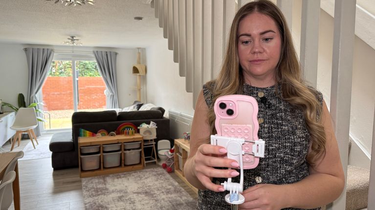 Nuori nainen seisoo olohuoneen ja portaiden vieressä edessään pinkki kännykkä.