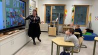 Opettaja luokan edessä osoittaa opetustaululle. Oppilaat nojaavat käsivarsiinsa. 