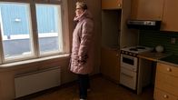Keskellä tyhjää vanhaa keittiötä, nainen katsoo ulkovaatteissaan ikkunasta ulos ja on sivuttain kohti kameraa.