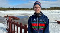 Kuvassa hiihtäjä hymyilee hiihtostadionilla. Yllään hänellä on kirjavat hiihtovaatteet ja silmälasit. Kaulassa on puffi-huivi.
