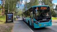 A light-blue bus at a stop with three people walking away and trees in the background.
