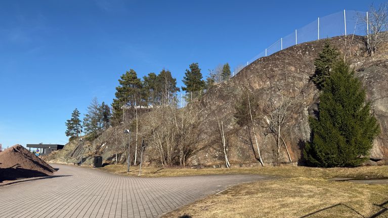 En stenlagd promenadväg, bredvid ser man ett högt berg och lite träd.