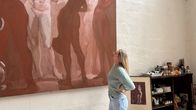 En kvinna står framför en stor målning, ser på tavlan som avbildar flera figurer. Vid sidan av finns en mindre inramad tavla lutad mot väggen.