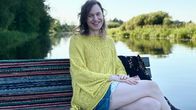 Keltapaitainen, pitkähiuksinen henkilö istuu penkillä, joka on laiturilla. Takana tyyni järvi ja vihreät puut.
