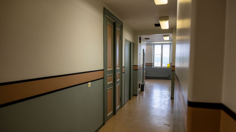 En tom korridor med grå och beige väggar, stängda dörrar och ett fönster i slutet av gången.