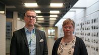 Två personer står inomhus i en hall med foton uppsatta längs ena väggen.