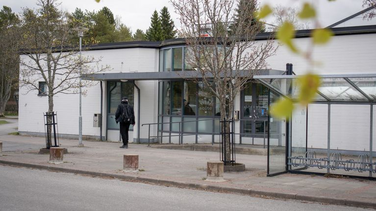 En person går mot ingången till en vit byggnad med cykelställ och träd i förgrunden.