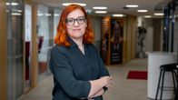 En person med axellångt rött hår står med armarna i kors i en kontorskorridor med glasväggar. 