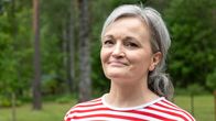  Harmaahiuksinen henkilö punavalkoraitaisessa paidassa seisoo ulkona vihreässä metsämaisemassa.