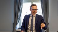 Mies istuu puupöydän ääressä toimistossa, pitää kädessään kansiota ja osoittaa pöydälle. Edessä on lasi vettä.