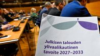 Taustalla ihmisiä palaverissa, etualalla paperinippu, jossa lukee Julkisen talouden ylivaalikautinen tavoite 2027-2033.
