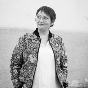 Toimittaja Kaisa Pulakka seisoo kevyesti hymyillen. Hänellä on lyhyet hiukset ja pyöreät silmälasit.