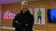 En person med svart tröja står med armarna i kors framför en vägg med affischer och en rosa neonskylt med texten "UMK".