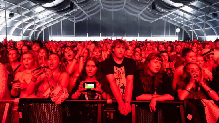 Konserttiyleisö odottaa innokkaana esityksen alkua suuressa tapahtumateltassa, valoissa punainen hehku.