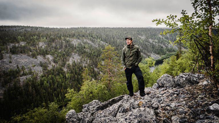 Mikko "Peltsi" Peltola seisoo korkealla kalliolla retkeilyvaatteet päällä. Taustalla sola ja havumetsää.
