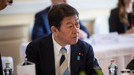 Pukuun pukeutunut mies istuu sisätilassa. Hänellä on raidallinen solmio kaulallaan. Pöydällä hänen edessään on pieni japanin lippu.