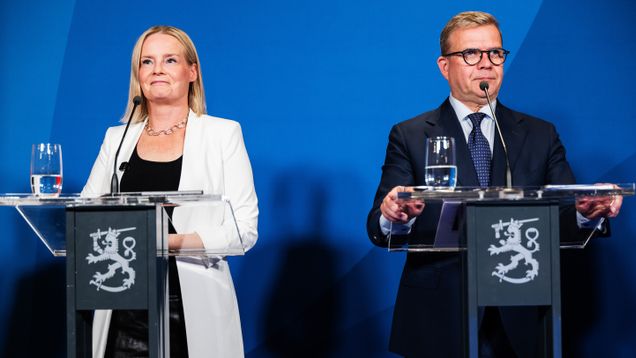 Riikka Purra i en svart blus och vit kavaj står bredvid en kostymklädd Petteri Orpo vid talarpodium.
