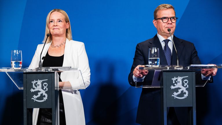 Riikka Purra ja Petteri Orpo seisovat puhujakorokkeilla sinisen taustan edessä, pöydillä lasilliset vettä ja mikrofoneja.