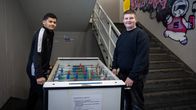 Two teens in dark casual clothing, standing by a foosball table, smiling at the camera.