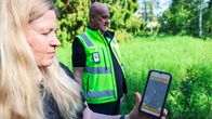 En person håller upp en mobil med karta synlig, ytterligare en person står bredvid i grön väst, utomhus i grön miljö.