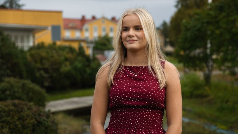 n ung person med ljust hår står utomhus framför buskar och gulmålade byggnader.