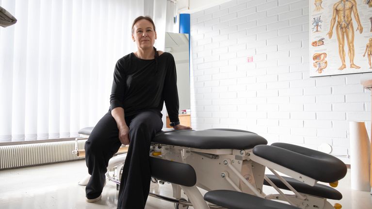 Henkilö istuu hoitopöydällä. Huoneessa on valkoinen tiiliseinä ja anatominen juliste, joka esittää lymfaattista järjestelmää.