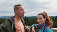 Mies ja nainen seisovat ja nojaavat kaiteeseen korkealla. Taustalla näkyy metsää, vaaroja ja pilvipoutaista taivasta.