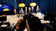 Sex personer sitter vid ett konferensbord framför blå gardiner och två stora blommor; texten “Kotimaa kutsuu” syns i bakgrunden.