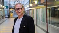 Silmälasipäinen mies seisoo kauppakeskuksen käytävällä ja katsoo kameraan.