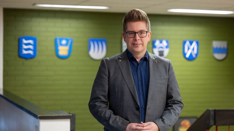Juha Herrala seisoo käytävällä ja katsoo kameraan. Takana seinällä on kunnan vaakunoita.
