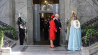 Sveriges kung  Carl XVI Gustaf iklädd mörk kostym och står tillsammans drottning Silvia vid en trappa. Silvia bär röd klänning och matchande hatt.