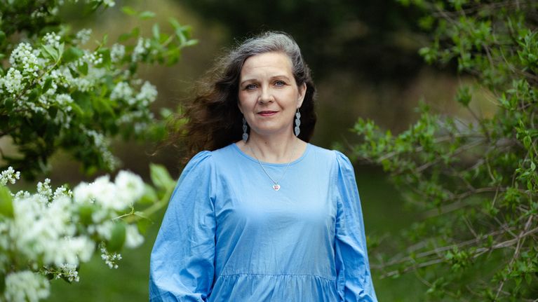 Teresa Nikkari, en av Svenska Yles Sommarpratare 2026, står i blå klänning utomhus bland gröna växter och vita blommor.