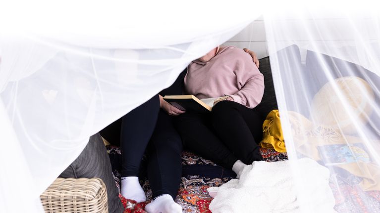 Kaksi henkilöä istuu sylitysten sohvalla värikkään peitteen päällä, toinen pitää avointa kirjaa sylissään. 