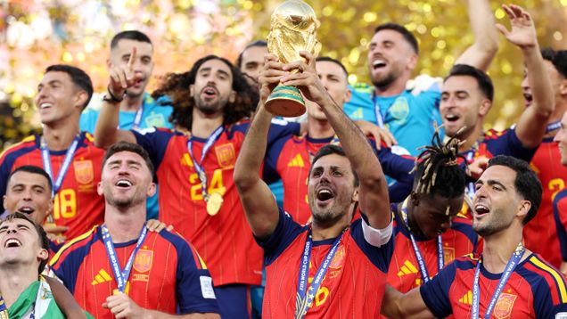 Members of the Spanish football team in red and blue jerseys celebrating their win, with one holding up the World Cup trophy.