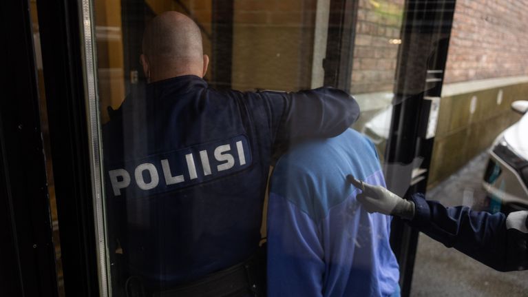 Poliisin virkailija saattaa sinitakkista henkilöä sisätiloihin osana virkatehtävää.