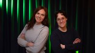 Två personer står bredvid varandra med armarna i kors framför ett mörkt draperi med gröna ljuseffekter.