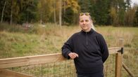 En person med ljust hår och svarta kläder står vid ett trästaket på en äng med träd i bakgrunden.
