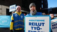 Ihmisiä banderollien kanssa talon edessä. ihmisen pitämässä taulussa lukee Ansaitsemme reilut työehdot.