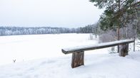 Lumipäällysteinen penkki maisemapaikalla. Taustalla näkyy jäätynyt järvi ja kaukainen metsänranta.