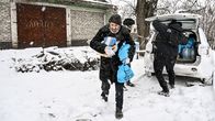 A few people in dark clothing collecting bags from the trunk of a car, snow is falling and on the ground.