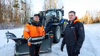 Två personer står framför en traktor med plog på en snötäckt väg. 