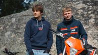 Två unga personer står utomhus framför en stor sten, intill en orange moped.